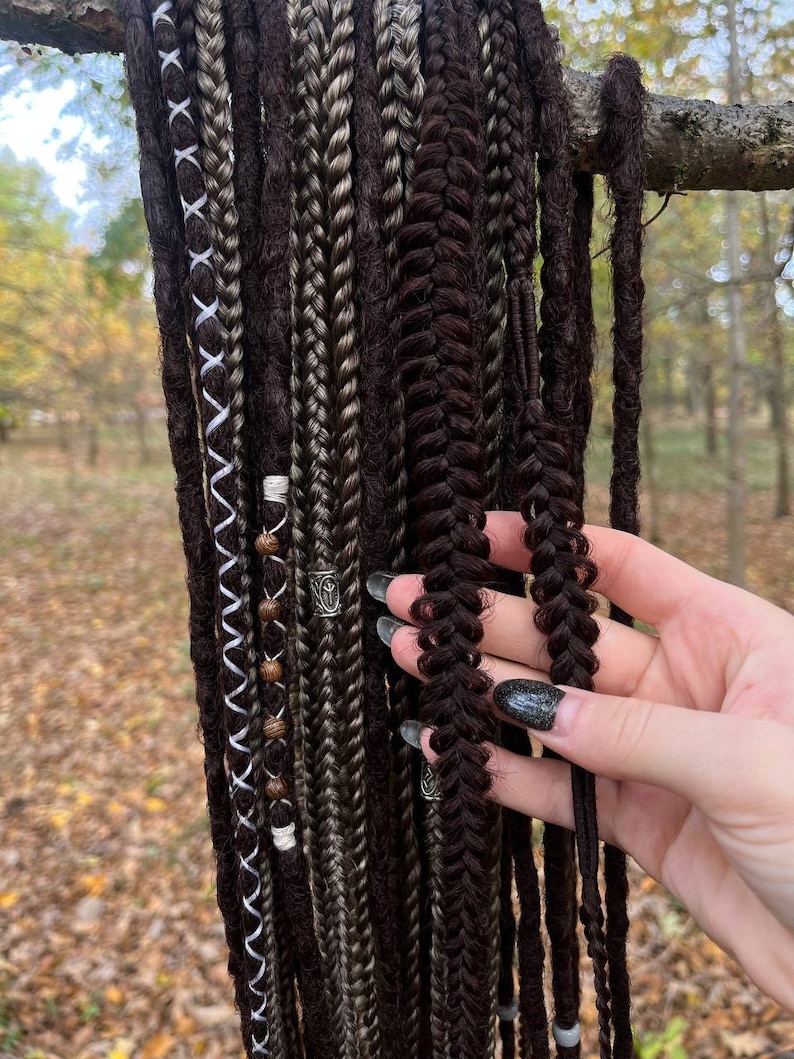 May include: A collection of brown, black and grey synthetic dreadlocks with beads and metal charms. The dreadlocks are hanging from a branch.
