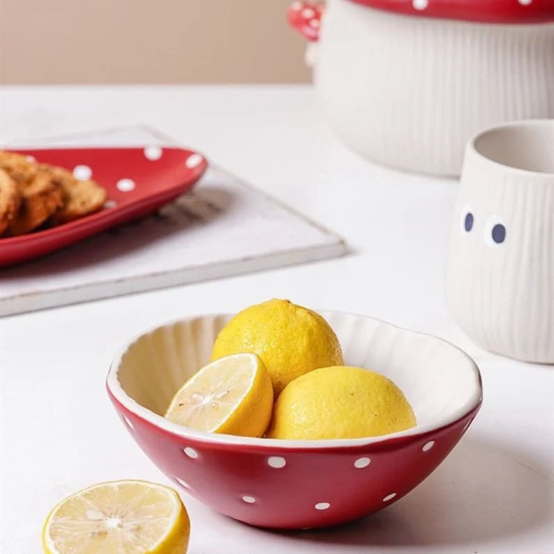 Pu&ograve; includere: Una ciotola in ceramica rossa con pois bianchi, contenente limoni. Sullo sfondo, un piatto rosso abbinato con biscotti e un contenitore bianco a forma di fungo. La ciotola ha l'interno bianco e il bordo smerlato.