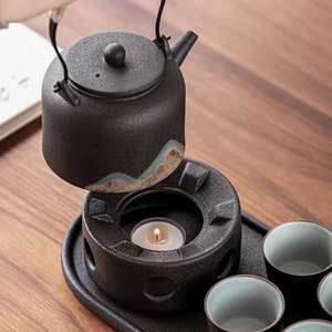 May include: A black ceramic teapot with a matching lid, a small spout, and a handle. The teapot sits above a black warmer with a lit candle. Several small teacups with blue interiors are also visible.
