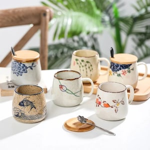 May include: A collection of ceramic mugs and a small jar with lids and spoons. The mugs feature hand-painted designs of flowers, birds, and fish in blue, red, and green on a cream background. Some mugs have wooden lids.