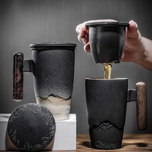 May include: Two black ceramic tea mugs with wooden handles and lids. One mug has a tea infuser. The mugs have a mountain design at the base. The tea is being poured from the infuser into the mug.