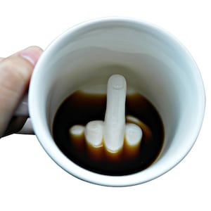 Può includere: Tazza da caffè in ceramica bianca con una scultura a forma di mano all'interno, che mostra il dito medio. La tazza è riempita di liquido scuro, probabilmente caffè o tè. La tazza è tenuta da una persona.