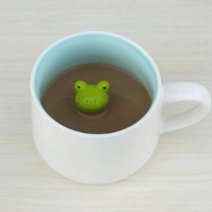 May include: A white ceramic mug with a light blue interior and a green frog-shaped lid floating in a brown liquid. The mug has a white handle and is filled with a brown beverage, possibly coffee or tea. The frog lid adds a whimsical touch.