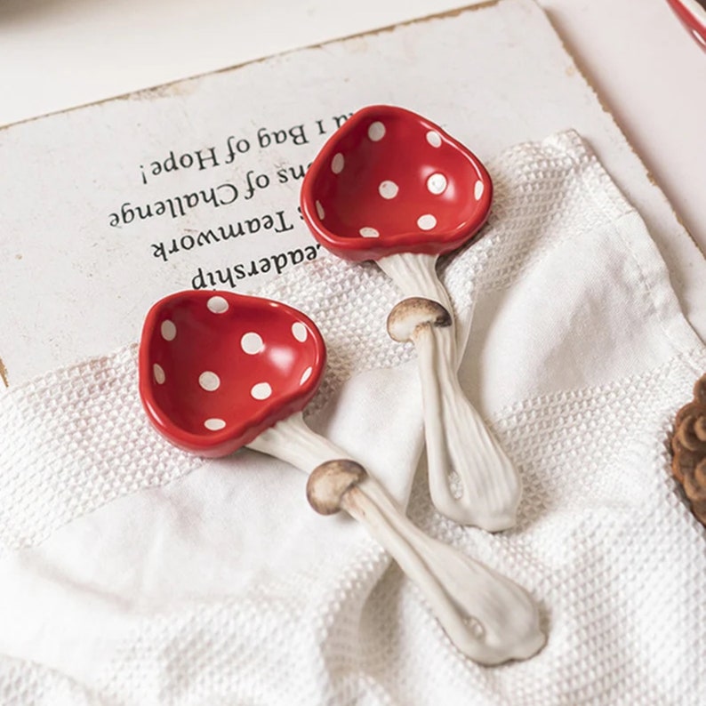 Pu&ograve; includere: Due cucchiai in ceramica a forma di fungo. Le teste dei cucchiai sono rosse con pois bianchi e i gambi sono bianchi con un dettaglio marrone a cappello. Sono appoggiati su una superficie bianca e strutturata. Cucchiaini decorativi e stravaganti.