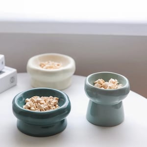May include: Three ceramic pet bowls in varying shades of green and cream, each filled with dry pet food. The bowls have a pedestal base and a rounded, tilted top. The bowls are arranged on a white surface.