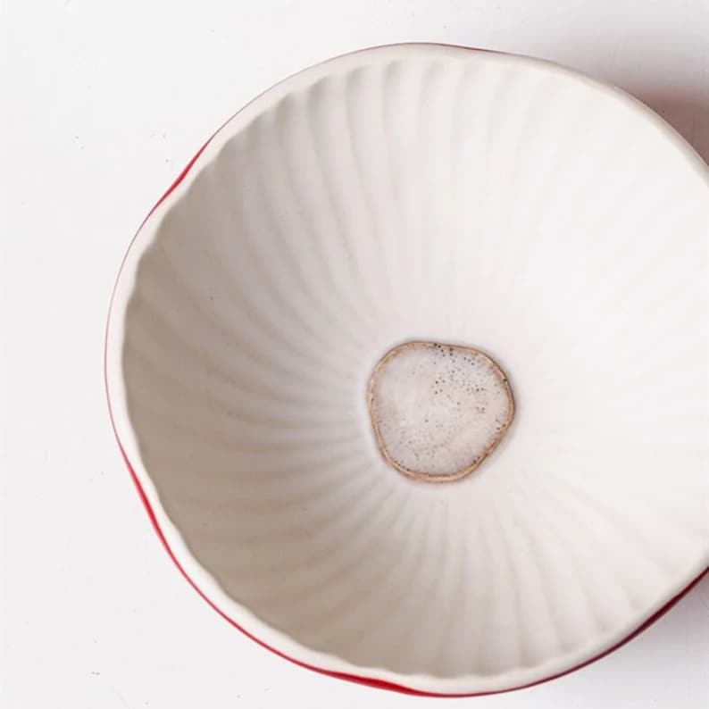 Pu&ograve; includere: Una ciotola in ceramica bianca con un motivo a spirale e un bordo rosso. Il centro della ciotola presenta una piccola pietra di colore chiaro di forma irregolare. La ciotola &egrave; probabilmente utilizzata per servire cibo o come elemento decorativo.