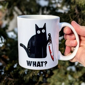 Op de afbeelding: Witte keramische mok met een illustratie van een zwarte kat die een bloedig mes vasthoudt. De kat heeft grote, ronde ogen en het woord "WHAT?" staat eronder. De mok heeft een wit oor.