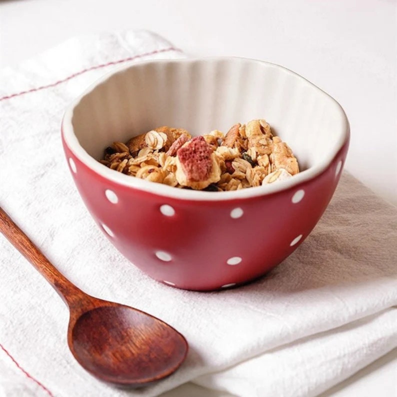 Pu&ograve; includere: Una ciotola di ceramica rossa con pois bianchi, piena di granola e frutta secca, &egrave; appoggiata su un panno bianco. Un cucchiaio di legno &egrave; accanto alla ciotola, pronto per una colazione sana.