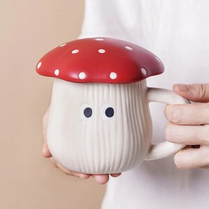 Pu&ograve; includere: Una tazza in ceramica stravagante a forma di fungo, con un cappello rosso a pois bianchi e un gambo color crema. La tazza ha un manico e due occhi a pois neri. La tazza &egrave; tenuta da una persona.