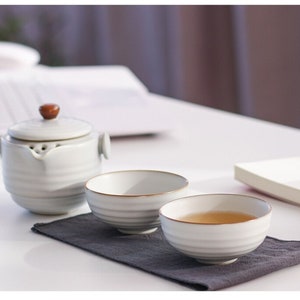May include: A white ceramic tea set comprising a teapot and two cups. The teapot features a brown wooden knob on the lid. The cups have a brown rim and are filled with tea. The set rests on a grey cloth.