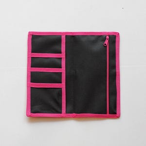 May include: Black organizer with a pink trim and zipper. The organizer has multiple pockets of varying sizes, including a mesh pocket. The trim and zipper are a vibrant pink, contrasting with the black fabric. The organizer is rectangular.