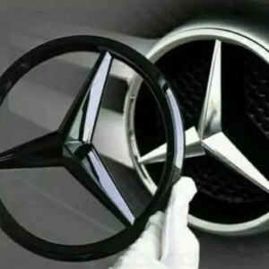 Op de afbeelding: Een zwart Mercedes-Benz embleem naast een zilveren embleem. Het zwarte embleem is een driepuntige ster in een cirkel. Het zilveren embleem is een driepuntige ster.