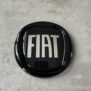 May include: A circular, glossy black Fiat emblem featuring the word "FIAT" in silver capital letters. The emblem has a ridged background. The image is taken from above, with the emblem centrally positioned.