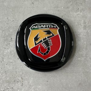 Könnte beinhalten: Ein schwarzes, kreisförmiges Emblem mit dem Wort "ABARTH" über einem Schild mit einem Skorpion-Design. Das Schild ist in gelbe, rote und weiße Abschnitte unterteilt. Das Emblem hat eine glänzende Oberfläche.