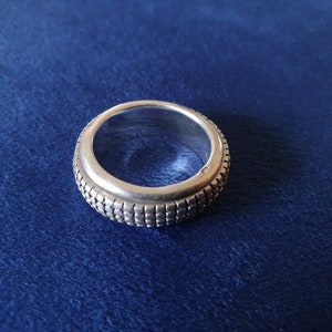 Peut inclure: Une bague en argent avec une bande texturée et à motifs. La bague est sur un fond bleu.