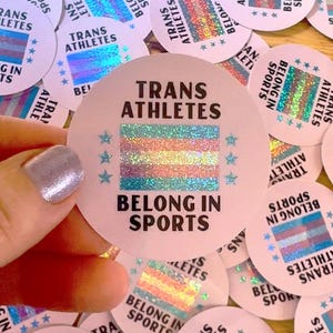 Peut inclure: Un gros plan sur un autocollant rond rose avec le texte "Trans Athletes Belong In Sports" en lettres noires. L'autocollant présente un drapeau de fierté transgenre holographique avec une finition scintillante.