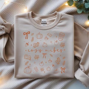 May include: A beige sweatshirt with the words "cozy vibes" in orange script, surrounded by orange illustrations of pumpkins, leaves, coffee cups, and bows. The Gildan brand label is visible at the top.