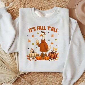 Könnte beinhalten: Hellblaues Sweatshirt mit dem Text "IT'S FALL Y'ALL". Die Grafik zeigt eine Gans mit braunem Pullover, die einen Kaffee hält und von Kürbissen und Herbstblättern umgeben ist. Ein herbstliches Design.
