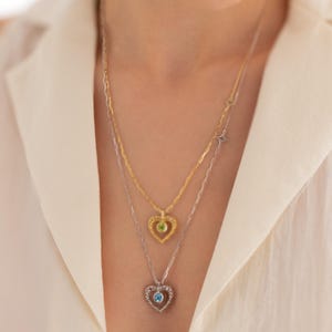 May include: Two delicate necklaces with heart-shaped pendants. The top necklace is gold with a green gemstone in the heart. The bottom necklace is silver with a blue gemstone in the heart.