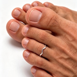 May include: A thin, polished silver-tone toe ring featuring a continuous, gentle wavy or serpentine design. The band is worn on the second toe, showcasing a smooth, reflective metallic surface that curves around the digit.