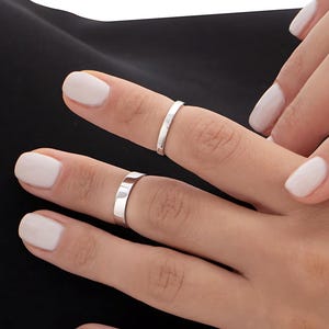 Könnte beinhalten: Zwei silberne Ringe an einer Hand mit weißem Nagellack. Die Ringe sind schlicht und dünn, mit einem flachen Band.