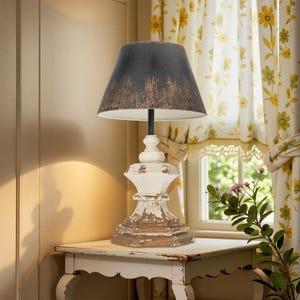 May include: A distressed white and brown table lamp with a black lampshade. The lamp has a weathered, antique appearance, with a wooden base and a textured lampshade. The lamp is placed on a white table.
