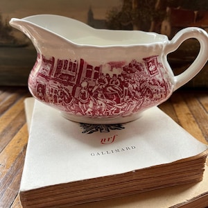 Peut inclure: Une saucière en céramique blanche et rouge avec une scène historique détaillée. Le bateau a un bec et une poignée. Il repose sur une pile de vieux livres, dont l'un porte le mot "GALLIMARD".