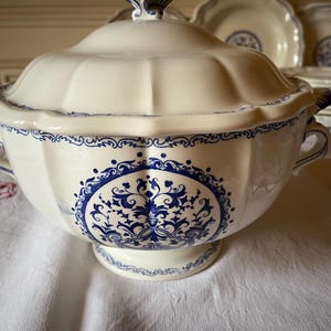 Peut inclure: Soupière en céramique crème avec un motif floral bleu et un couvercle assorti. La soupière a un bord festonné et deux poignées. Des assiettes assorties sont visibles en arrière-plan.