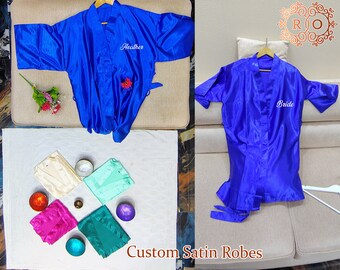 May include: Two blue satin robes with personalised embroidery. One robe says "Heather" and the other says "Bride". The robes are laid out on a white surface with other robes in different colours.