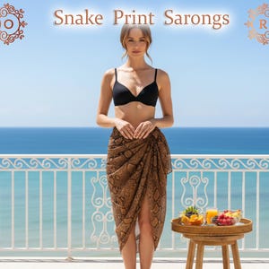 May include: A woman wears a brown snake print sarong and a black bikini top on a balcony with an ocean view. The image displays the text "Snake Print Sarongs". A small table holds fruit and juice. The sarong is a beachwear item.
