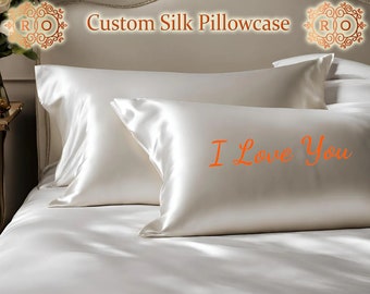May include: Two white silk pillowcases, one with the words "I Love You" embroidered in orange on the front. The pillowcases are on a white bed with a gold headboard.