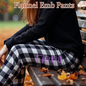 May include: Black and white checked flannel trousers with the name "Fiona" embroidered on the front. The trousers are being worn by a person sitting on a wooden bench with autumn leaves on the ground.