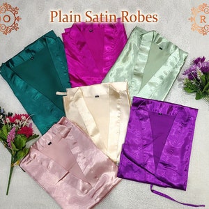 May include: Six satin robes in different colors, including green, pink, beige, light green, purple, and pink. The robes are laid out on a white surface. The text "Plain Satin Robes" is visible at the top of the image.