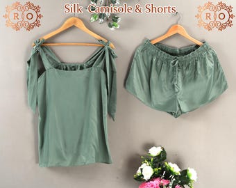 May include: A sage green camisole and shorts set featuring the text Silk Camisole & Shorts. The camisole has wide shoulder straps that tie into bows, while the shorts feature an elasticated waistband with a drawstring tie, both on wooden hangers.