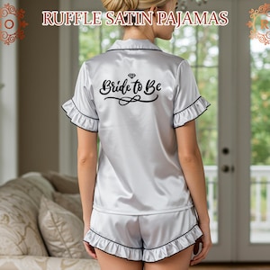 May include: White satin pajamas with black trim and ruffles. The top has short sleeves and the text "Bride to Be" with a diamond graphic printed on the back.