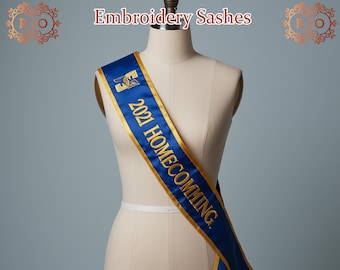 Op de afbeelding: Een blauwe satijnen sjerp met gouden rand en goud geborduurde tekst '2021 HOMECOMING'. De sjerp is over een mannequin gedrapeerd. De woorden 'Embroidery Sashes' staan bovenaan de afbeelding.