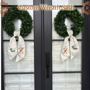 May include: Two wreaths with white satin sashes, each with a gold letter Z and a colourful masquerade mask embroidered on the sash. The wreaths are hung on a black door with glass panels. The text "Custom Wreath Sash" is displayed above the wreaths.