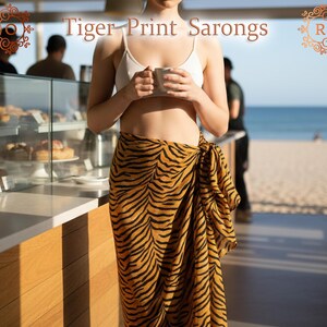 Può includere: Una donna indossa un sarong con stampa tigrata, con un top bianco, che tiene una tazza. Il sarong presenta un motivo a strisce nere su sfondo marrone. Il testo "Tiger Print Sarongs" è visualizzato nella parte superiore dell'immagine.