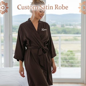 Peut inclure: Robe en satin marron avec une ceinture assortie. La robe a un col en V et des manches trois-quarts. Le texte "Custom Satin Robe" est en haut de l'image. Le nom "Lydia" est brodé sur la poitrine gauche.