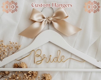 Könnte beinhalten: Ein weißer Holz Kleiderbügel mit einer goldenen Draht-Inschrift "Bride". Der Kleiderbügel ist mit einer beigefarbenen Schleife aus Satin verziert.
