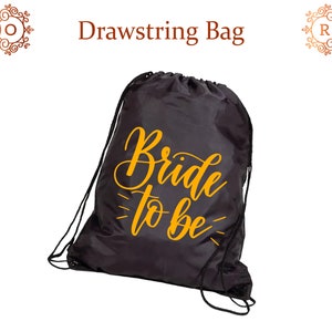 May include: Black drawstring bag featuring the words "Bride to be" in gold script. The bag has black drawstrings and is set against a white background. A practical and stylish accessory.