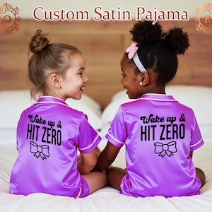 May include: Two girls wearing matching purple satin pajamas with short sleeves and white piping. The pajamas have a black graphic that says "Wake up & HIT ZERO" with a black bow underneath the text.