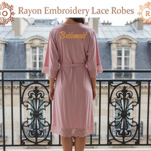 May include: A dusky pink rayon robe featuring lace detailing and a matching belt. The word "Bridesmaid" is embroidered in gold on the back. The robe has three-quarter sleeves and is knee-length. The text "Rayon Embroidery Lace Robes" is at the top.