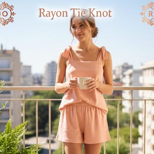 Può includere: Un completo pigiama annodato in rayon color pesca. Il set comprende una canotta con spalline annodate e pantaloncini abbinati. La modella tiene in mano una tazza bianca. Il testo "Rayon Tie Knot" è visualizzato nella parte superiore dell'immagine.