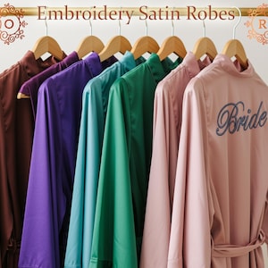 Op de afbeelding: Een verzameling satijnen badjassen in verschillende kleuren, waaronder bruin, paars, blauwgroen, groen en roze, hangend aan houten kleerhangers. De badjassen worden getoond op een gouden stang. E&eacute;n roze badjas heeft het woord "Bride" geborduurd.