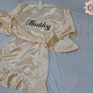 May include: A cream-colored satin robe with a ruffle trim and a black embroidered name "Maddog".