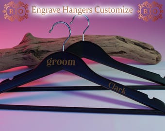 May include: Two black wooden hangers with silver hooks. One hanger is engraved with the word "groom" in gold, and the other with the name "Clark". The image includes the text "Engrave Hangers Customize".