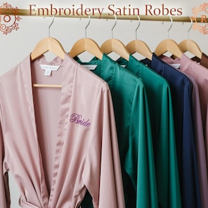 Könnte beinhalten: Eine Reihe von Satin-Roben in verschiedenen Farben, darunter Rosa, Grün und Marineblau, hängen an Holzbügeln. Auf einer der rosa Roben ist das Wort "Bride" in lila Schrift gestickt. Der Text "Embroidery Satin Robes" ist oben zu sehen.