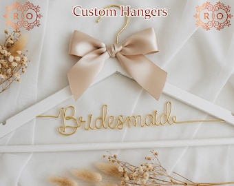 Könnte beinhalten: Ein weißer Holz Kleiderbügel mit einer goldenen Drahtinschrift, die "bridesmaid" lautet. Der Kleiderbügel ist mit einer beigefarbenen Schleife aus Satin verziert.