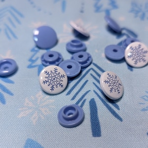 May include: A collection of blue and white plastic buttons with a snowflake design. The buttons are scattered on a blue fabric background with a snowflake pattern.